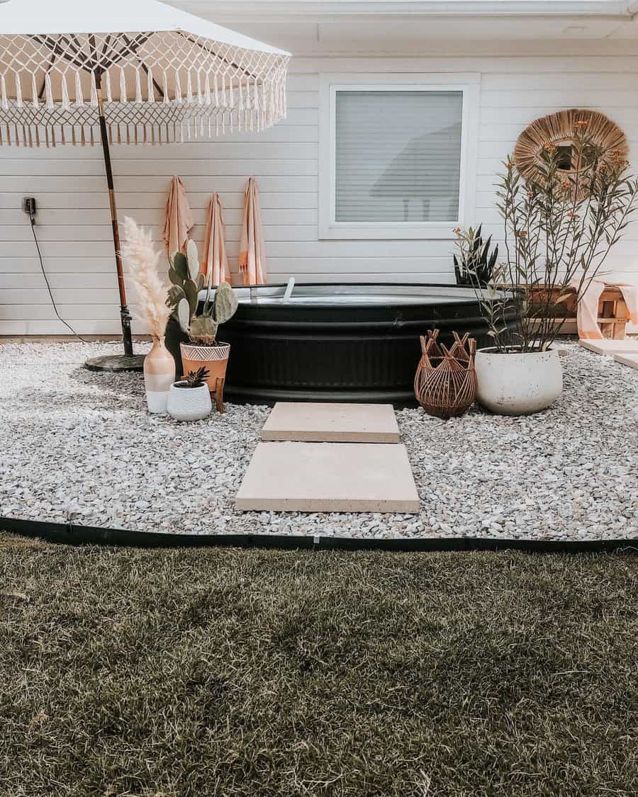 Small backyard with a round pool, umbrella, and potted plants on gravel, grass in the foreground and a house wall in the background