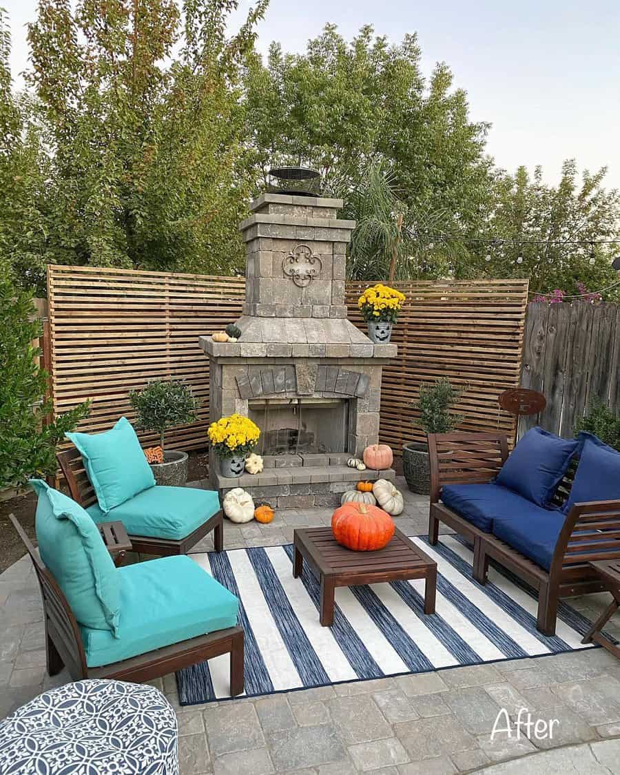 Cozy patio with a stone fireplace, pumpkins, potted plants, and blue cushioned wooden chairs around a striped rug