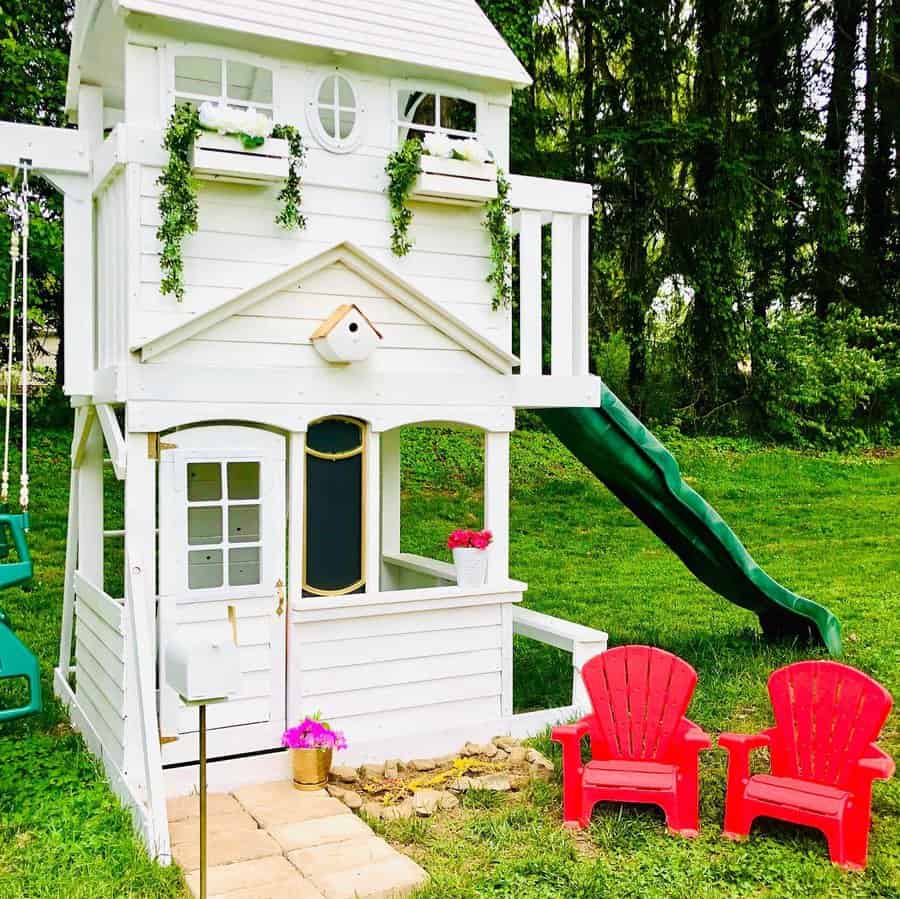 White two-story backyard playhouse with a green slide, swing, and two red chairs, surrounded by trees and greenery