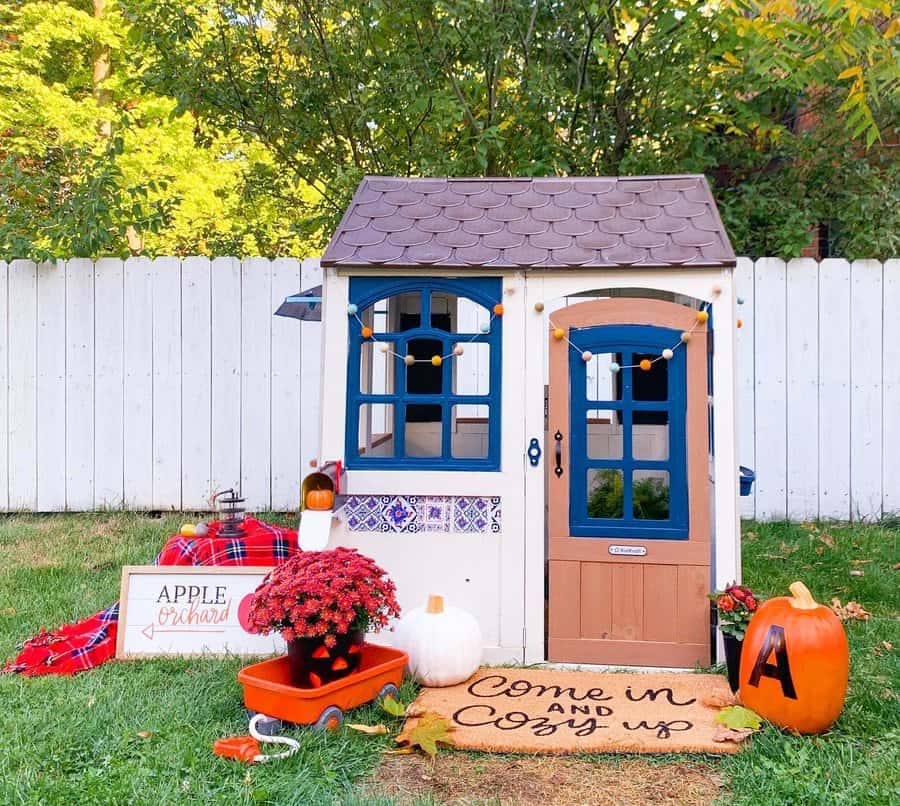 A small playhouse with fall decorations, pumpkins, a welcome mat, flowers, and a red wagon, set against a white fence and trees