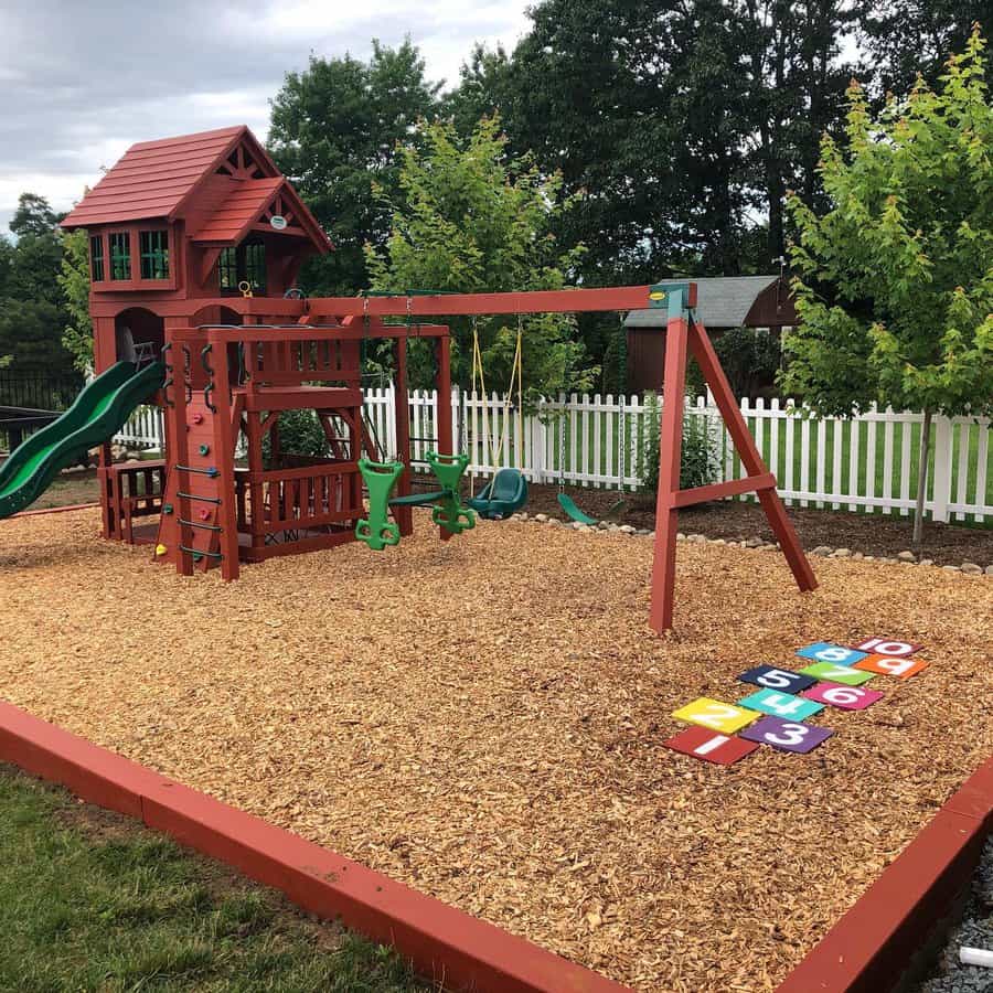 A backyard playground set with a slide, swings, climbing wall, and hopscotch on wood chips, surrounded by a white fence and trees