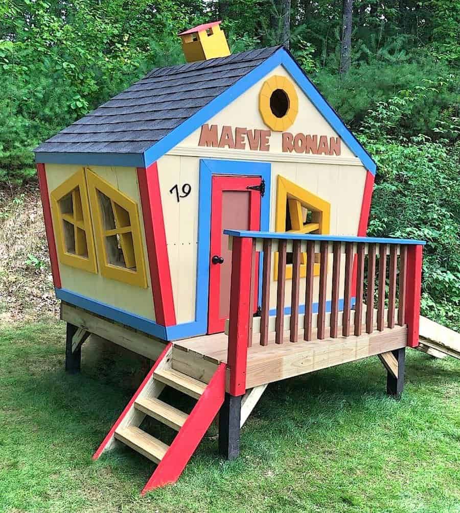 Colorful playhouse with a slanted roof, small porch, and bright red door, set on stilts with steps, surrounded by greenery