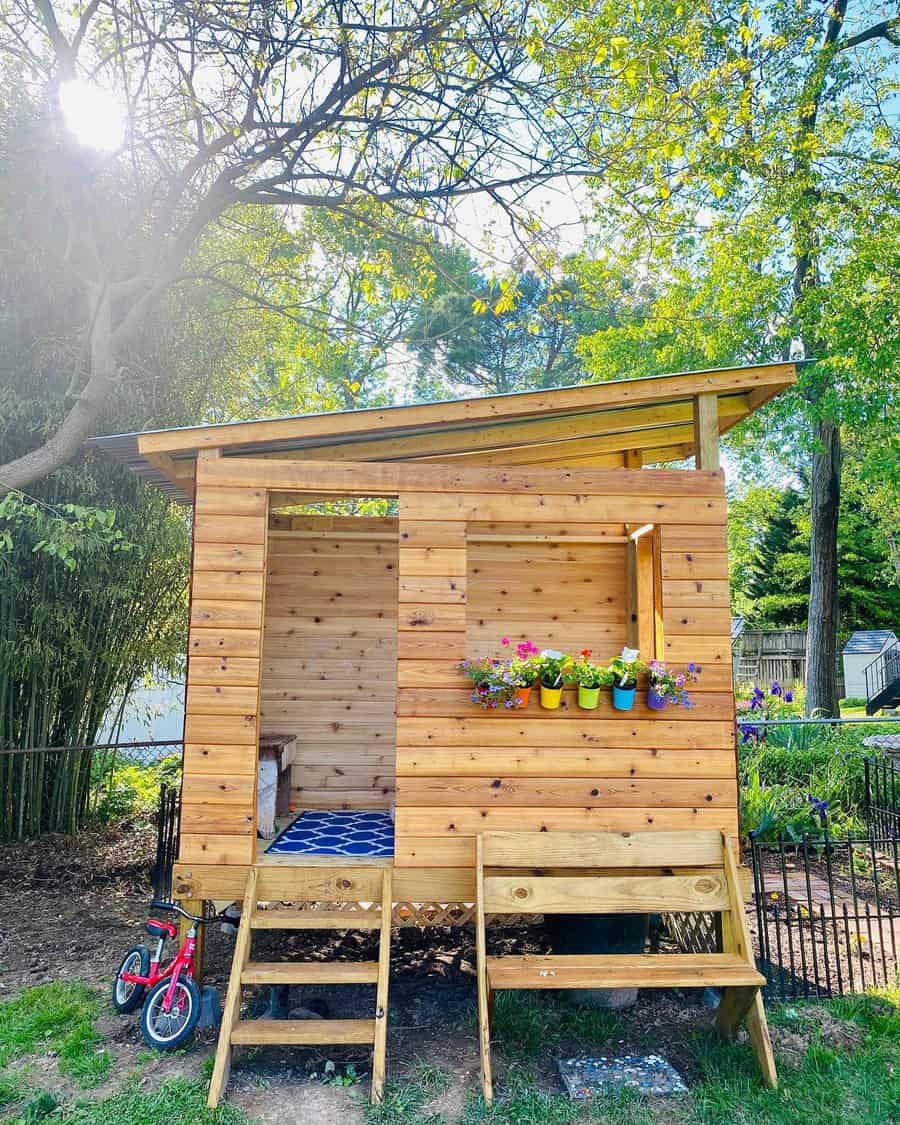 A small wooden playhouse with colorful flower pots on the window ledge, a small red bike nearby, and trees in the background