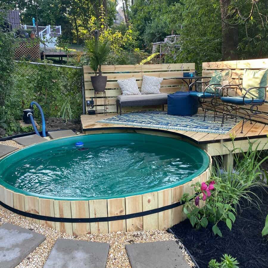 A small, round wooden pool with clear water, surrounded by a deck with seating, cushions, and plants in a lush garden setting