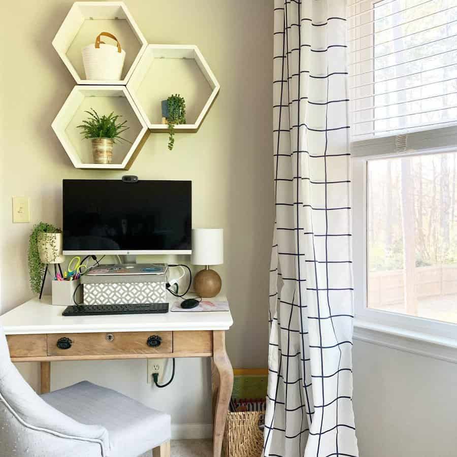 A home office corner with a wooden desk, chair, computer, and hexagonal shelves holding plants and decor. A window with grid-patterned curtains