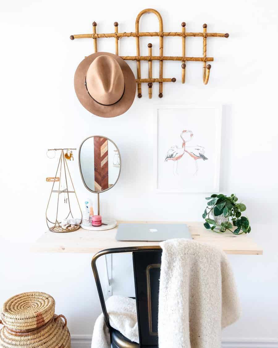 Minimalist desk with a laptop, plant, and decor; wall-mounted hat rack with a hat, and artwork; cozy chair with a beige throw