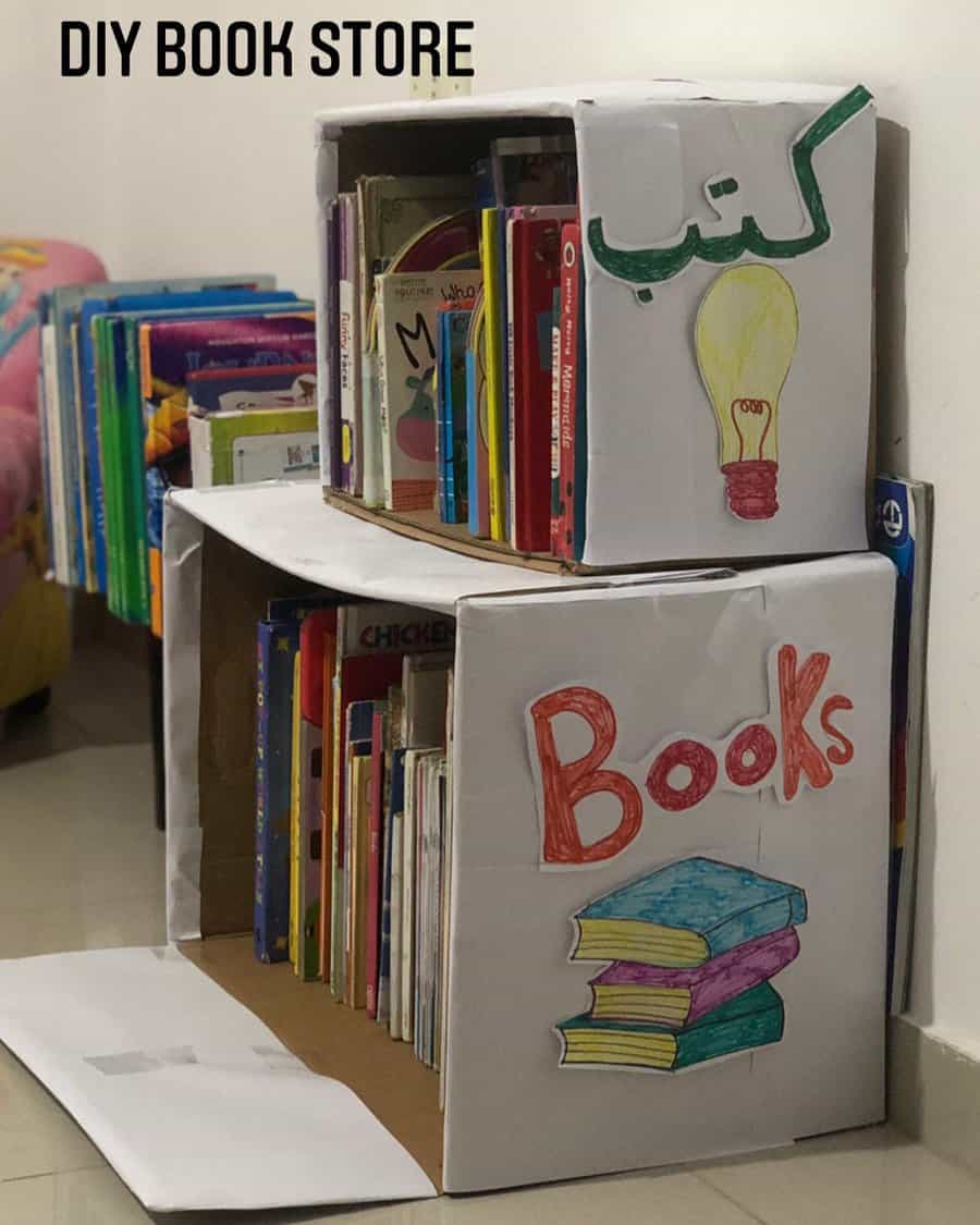 DIY book storage made from cardboard boxes, decorated with hand-drawn labels and filled with colorful children's books, placed on the floor.