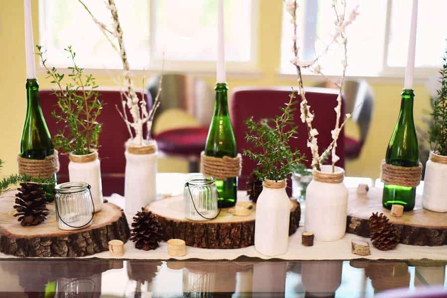 Rustic centerpiece with green bottles, white jars, candles, and pinecones on a table