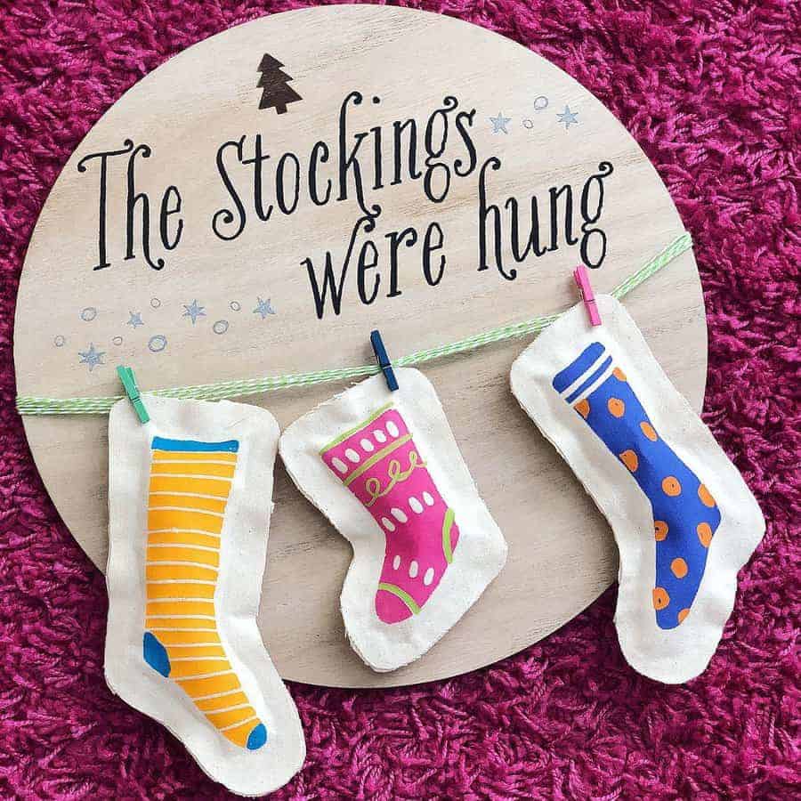 Three colorful stockings hung on a wooden sign with text "The Stockings Were Hung"