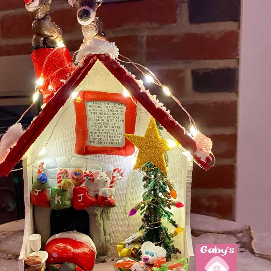 A festive holiday scene with a small, decorated house, reindeer figurines, stockings, a Christmas tree, and twinkling lights