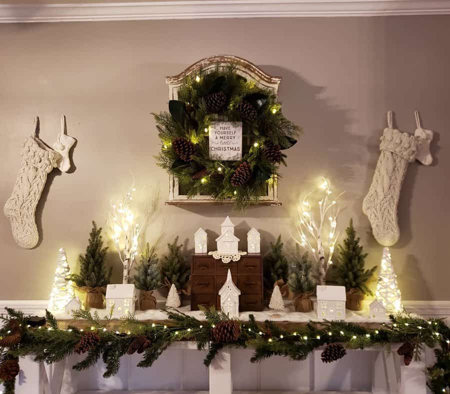 Festive mantel with two stockings, a wreath, mini Christmas trees, illuminated decor, and a small village display