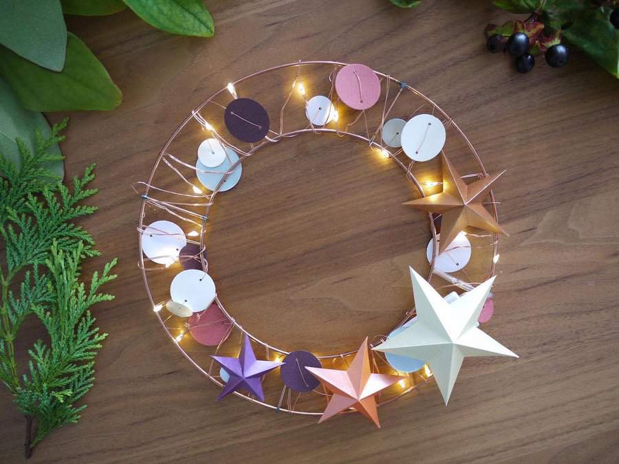 Decorative wreath with paper stars and fairy lights on a wooden table, surrounded by greenery and berries