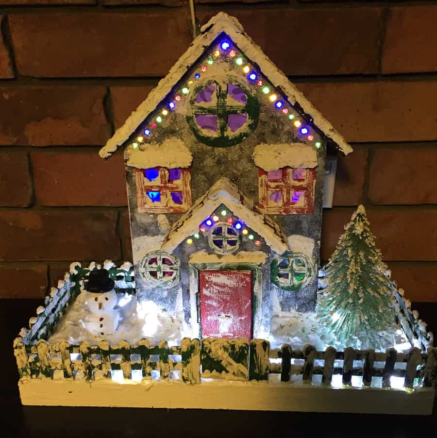 A small, illuminated, snow-covered cardboard house with a red door, colorful lights, a snowman, and a tree in a fenced yard