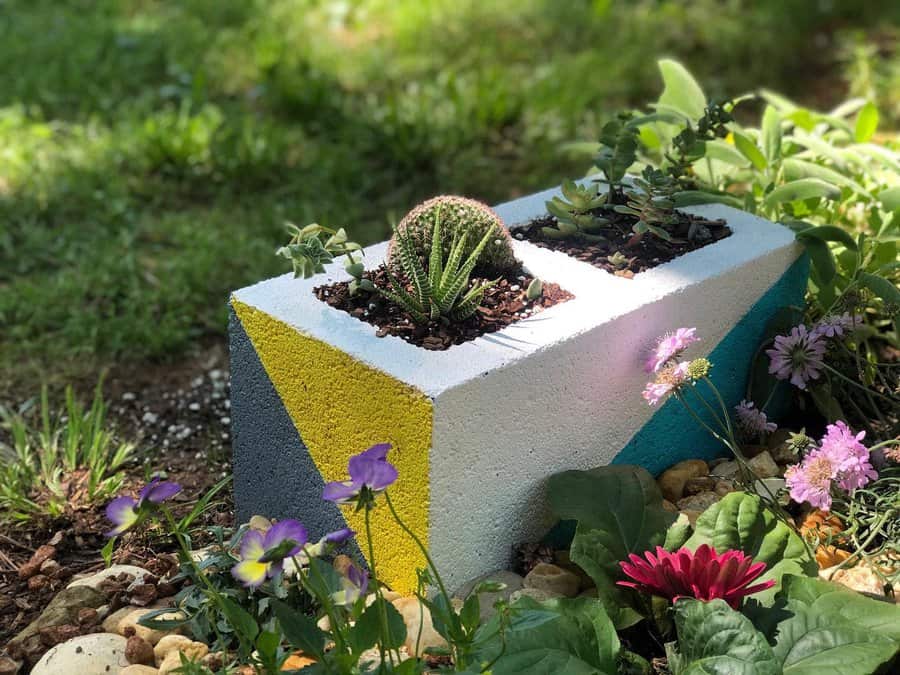 Colorful DIY cinder block planter with geometric yellow, blue, and white accents, filled with succulents and cacti, adding charm to a garden setting