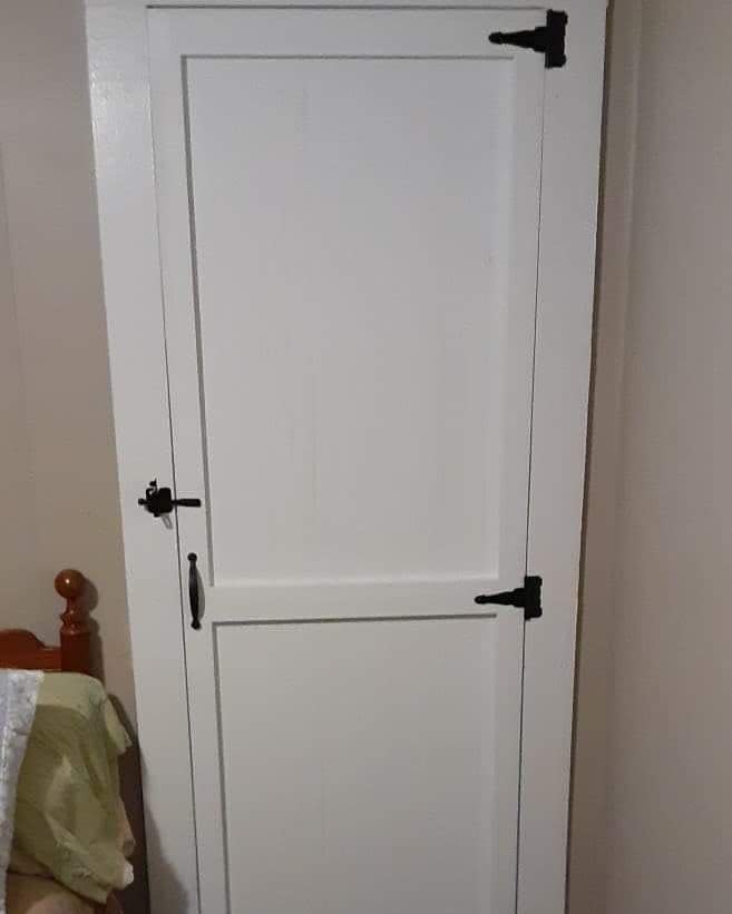 A white wooden door with black metal hinges and handles, adjacent to a bed with a green and white blanket