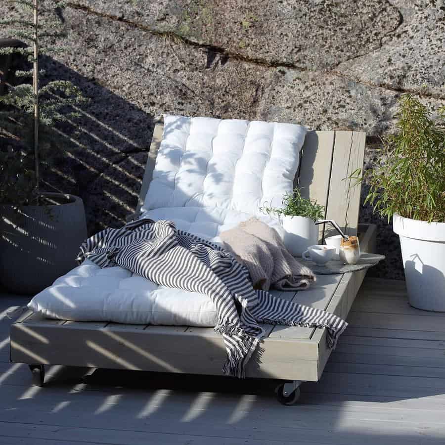 Outdoor daybed with white cushions, striped and beige blankets, a mug, and a potted plant on a patio against a stone wall backdrop