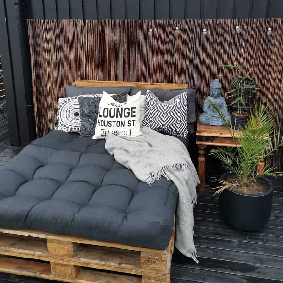 Rustic DIY pallet daybed with a plush black cushion, cozy pillows, and a bamboo privacy screen, creating a relaxing outdoor retreat