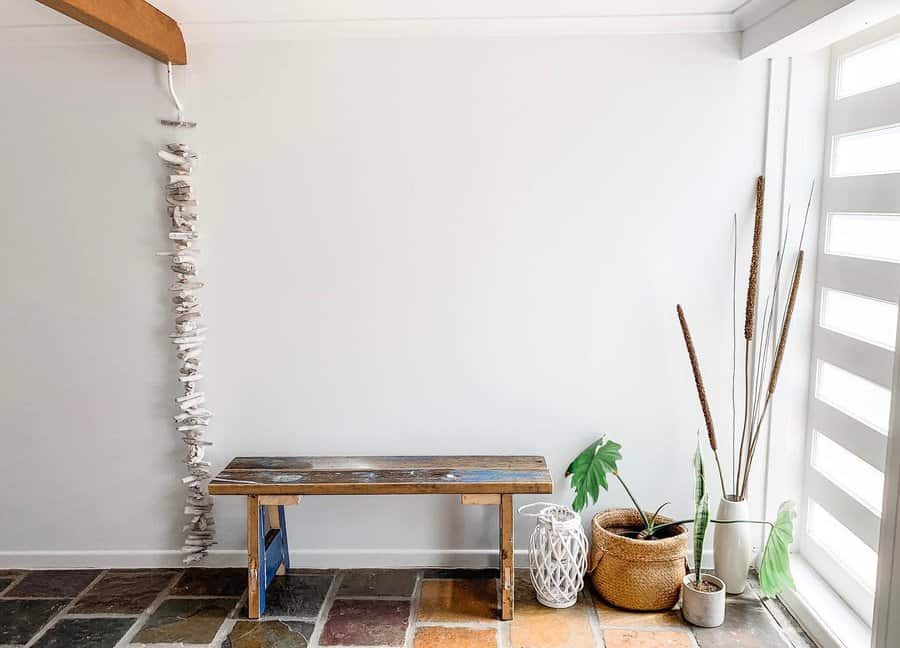 Minimalist entryway with a wooden bench, hanging driftwood, potted plants, and light filtering through striped blinds on the right