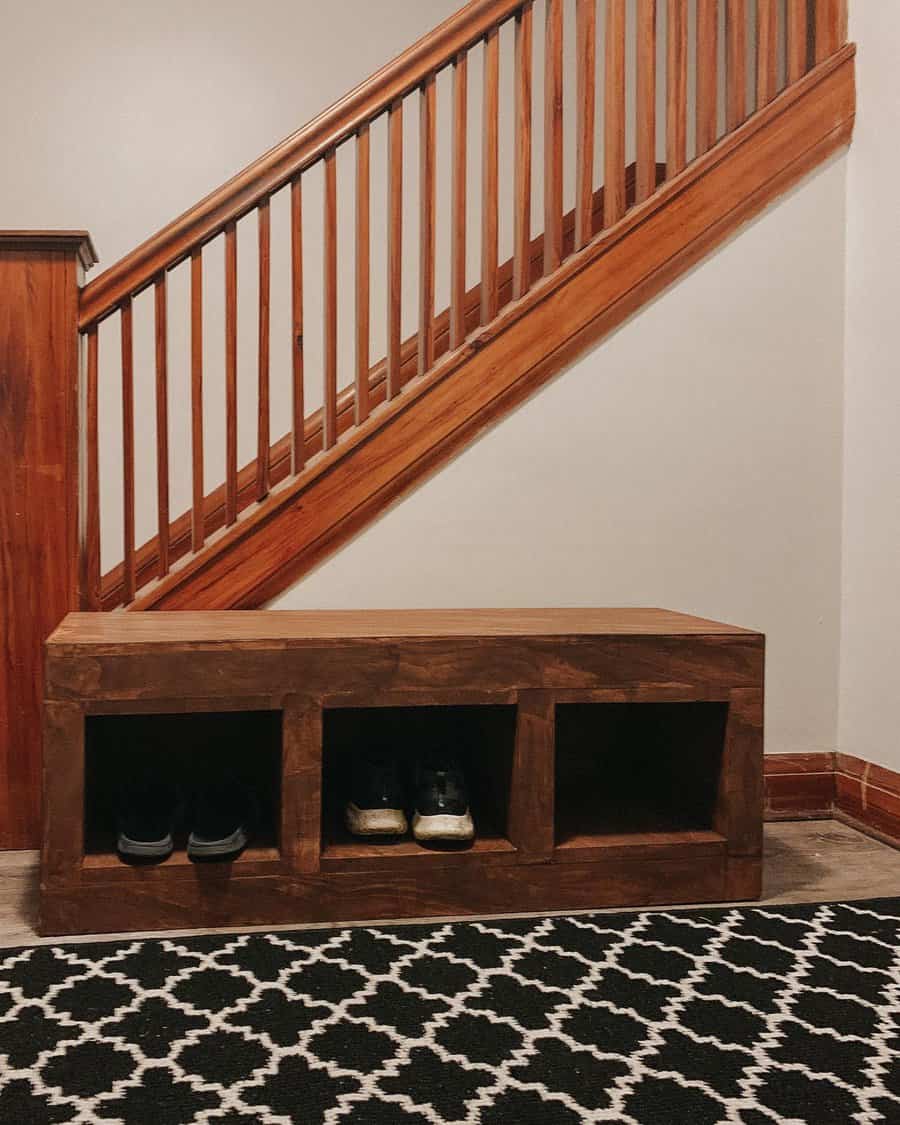 Wooden bench with cubbies holding shoes, placed under a wooden staircase, black and white patterned rug on the floor