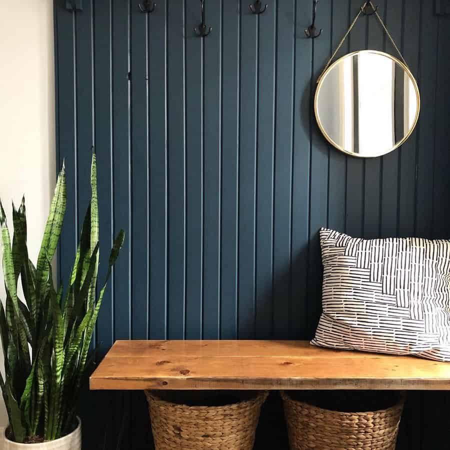 Entryway with a wooden bench, striped pillow, two woven baskets, a round mirror, and a snake plant against a dark blue wall