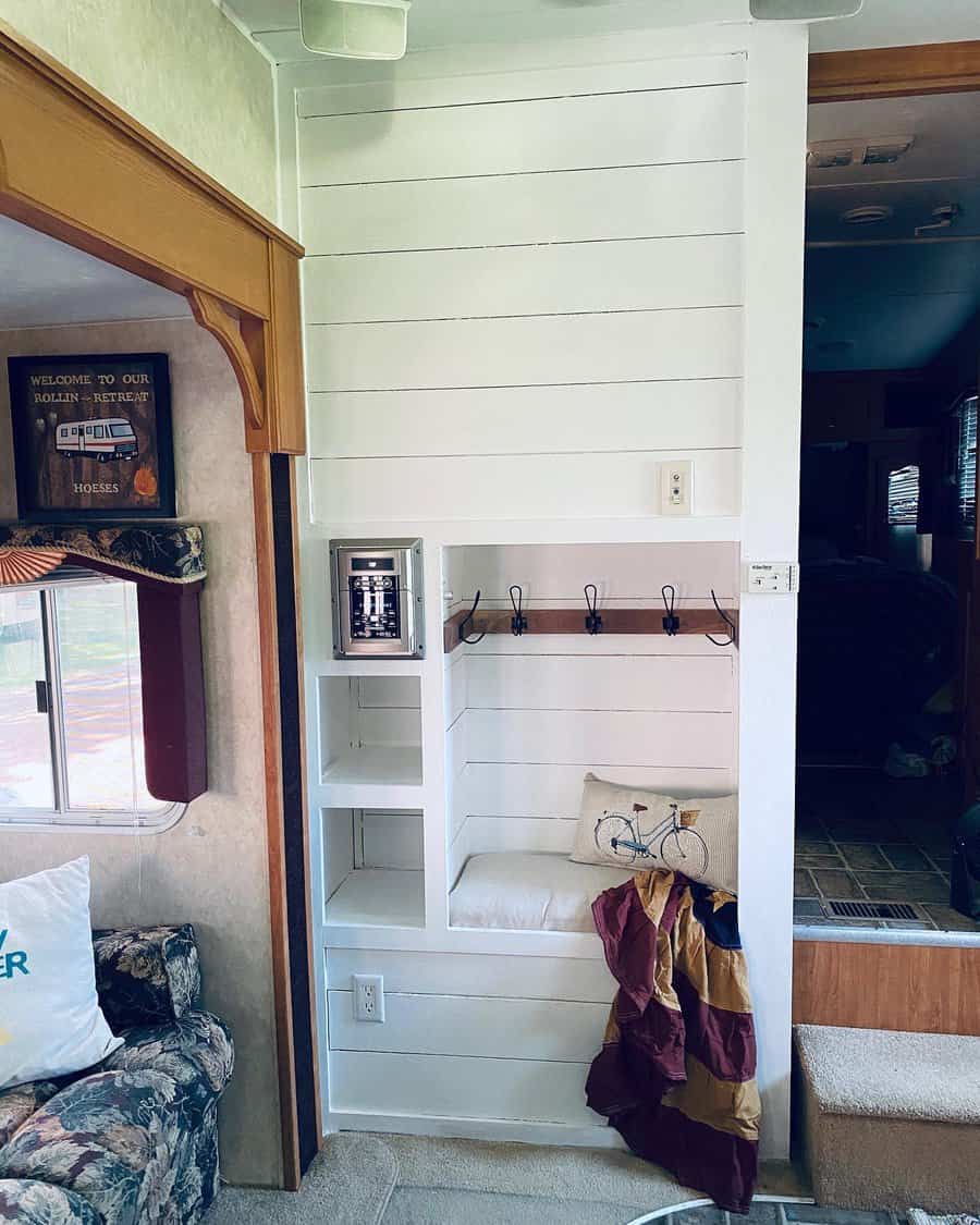 Compact RV interior with white shelves, hooks, a small bench with a bicycle cushion, and a wall outlet