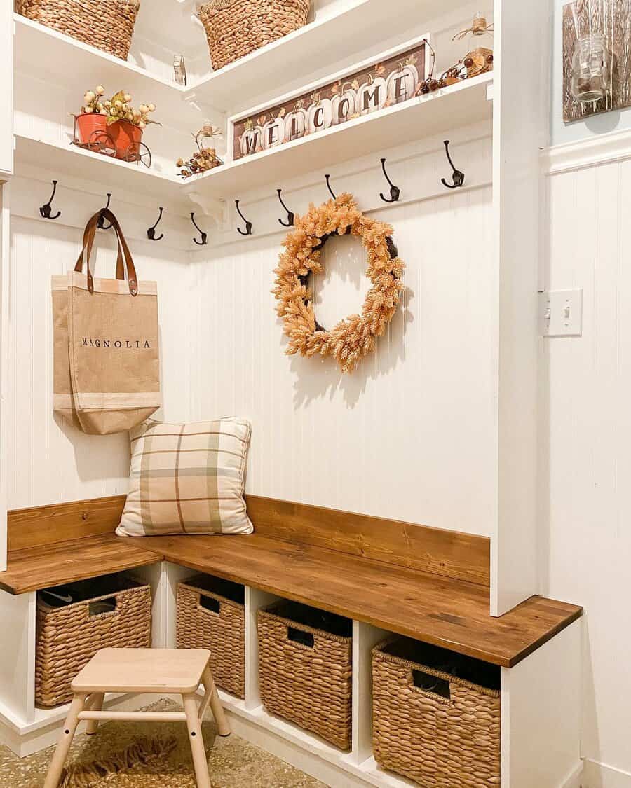 Cozy entryway with a wooden bench, woven baskets, hooks, plaid pillow, wreath, welcome sign, and a small stool