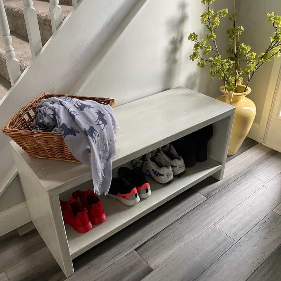 Shoe bench with red and white sneakers, a wicker basket with a blanket, and a yellow vase with branches, beside a staircase