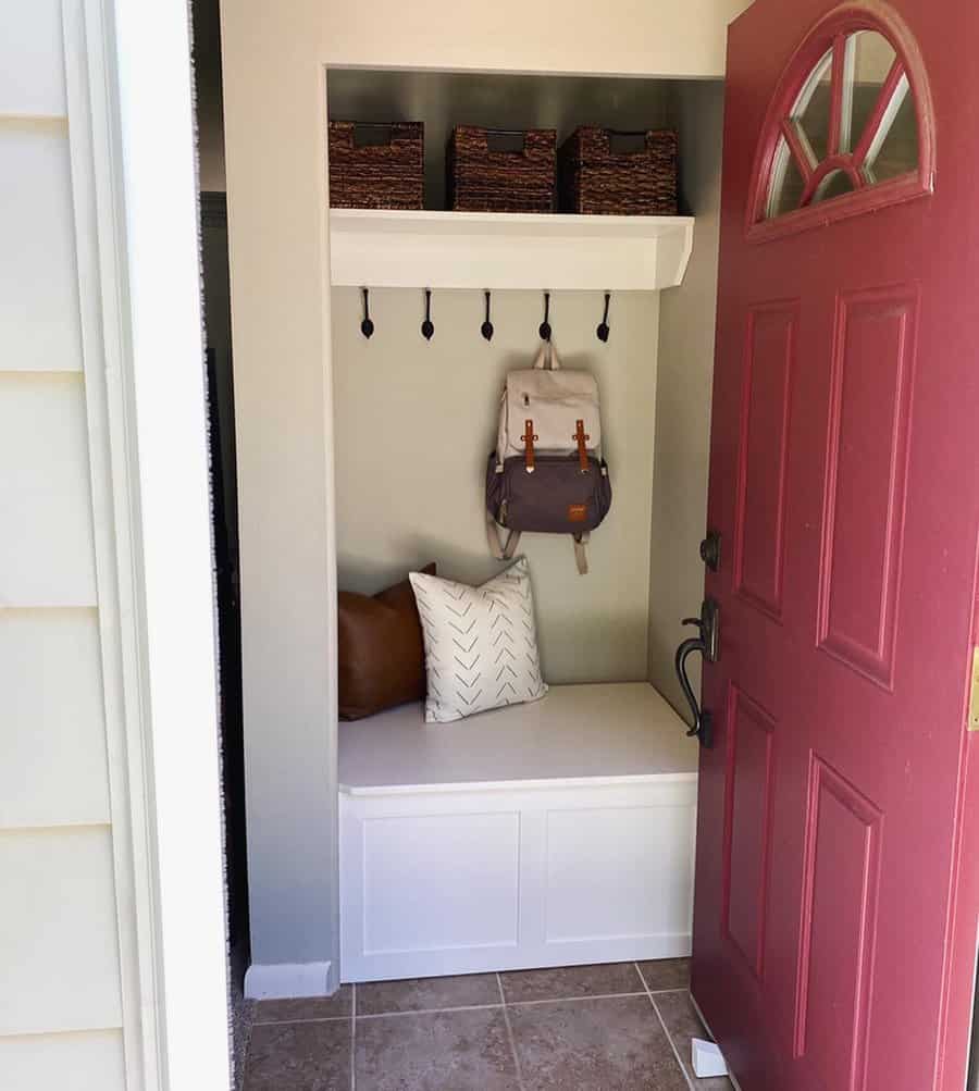 Entryway with an open red door, showing a white bench with pillows, hooks holding a backpack, and shelves with woven baskets above