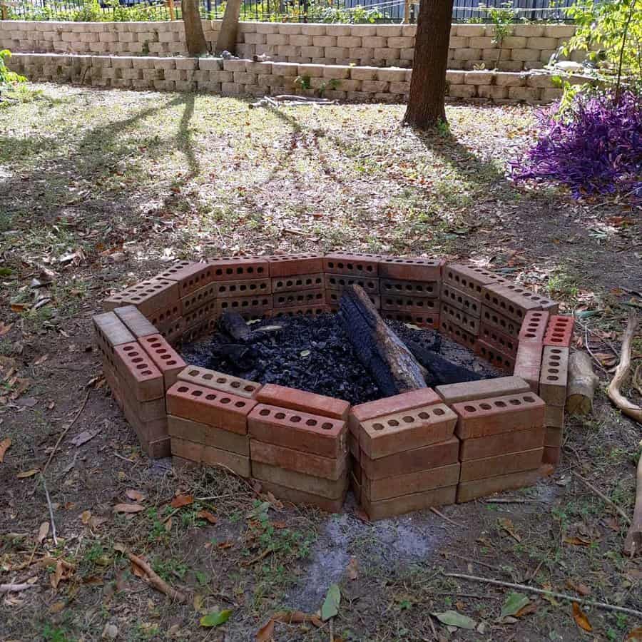 Brick fire pit with charred logs in a grassy area surrounded by trees and fallen leaves