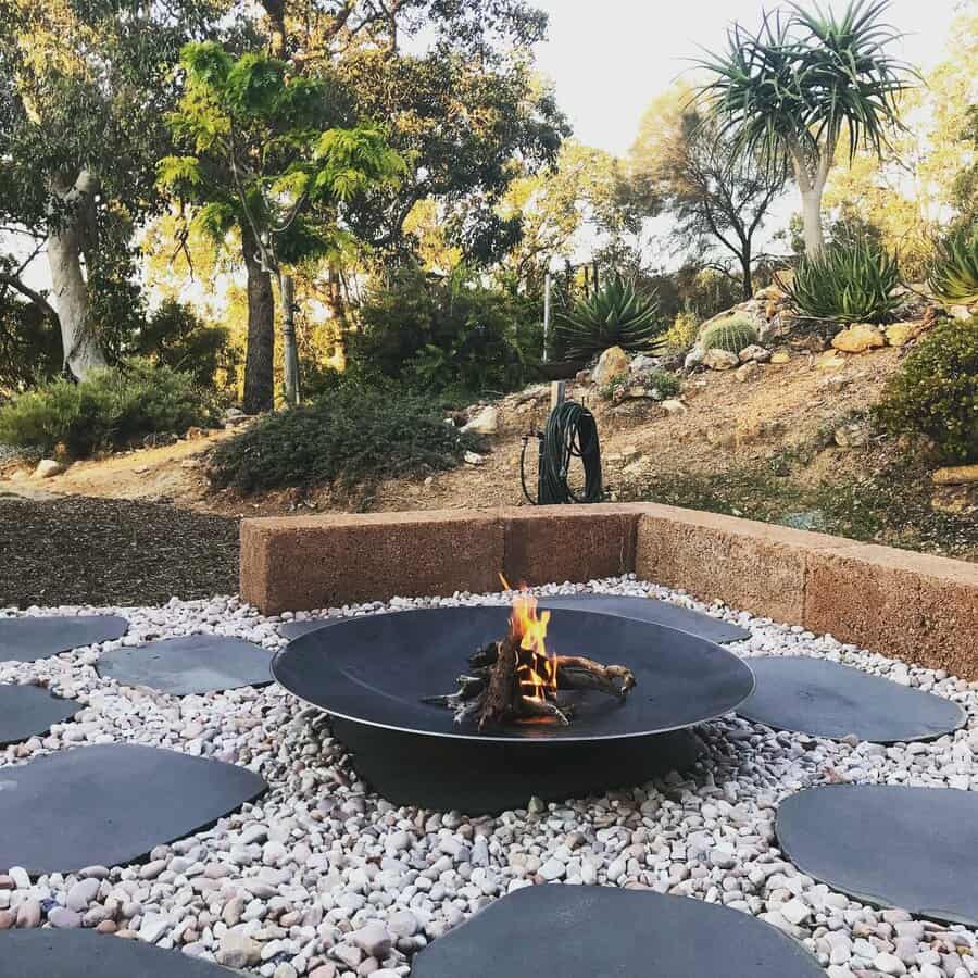 A black fire pit with a small flame sits on a gravel patio surrounded by stepping stones, with trees and plants in the background