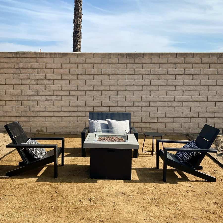 Outdoor seating with three black chairs around a rectangular fire pit on sandy ground, set against a brick wall and clear sky