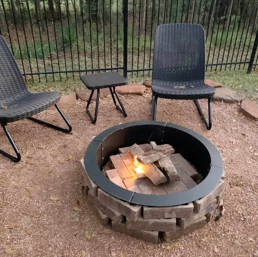 Outdoor fire pit with a small flame, surrounded by wooden logs; two wicker chairs and a small table nearby; fenced yard in the background