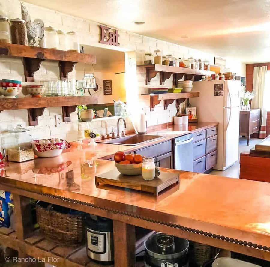 Cozy kitchen with copper countertops, wooden floating shelves stocked with jars, a white fridge, and a basket of apples on the counter