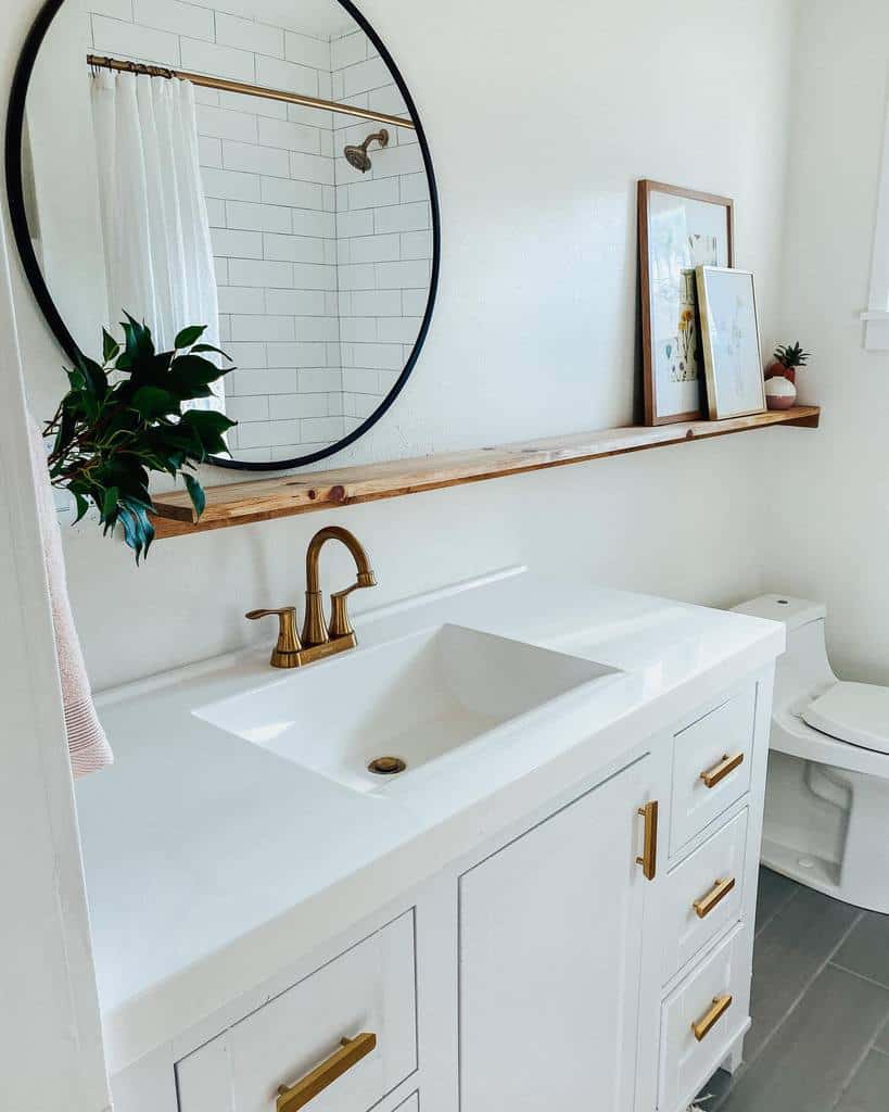 Minimalist bathroom with a white vanity, gold faucet, round mirror, and shelf with decor above, white tiled shower in the background