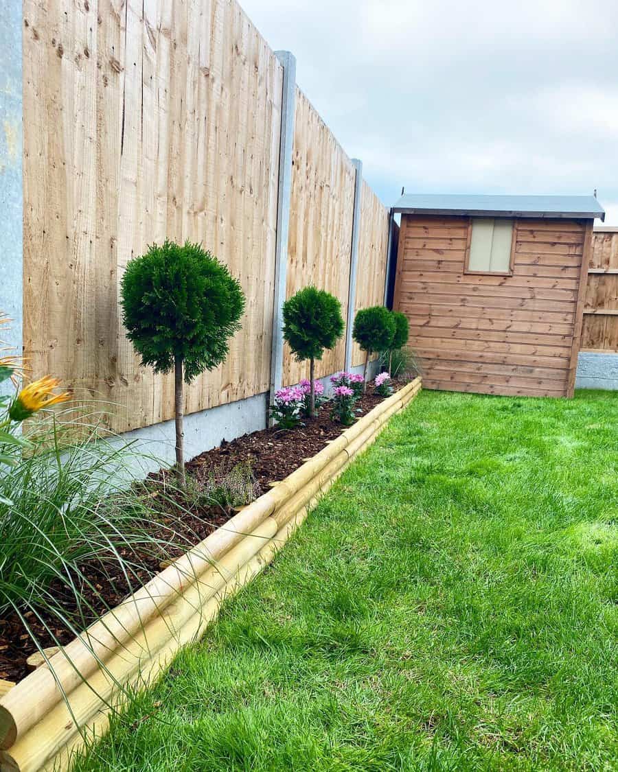 Simple backyard with wooden fencing, a small garden bed with topiary trees and flowers, and a wooden shed on a lush green lawn