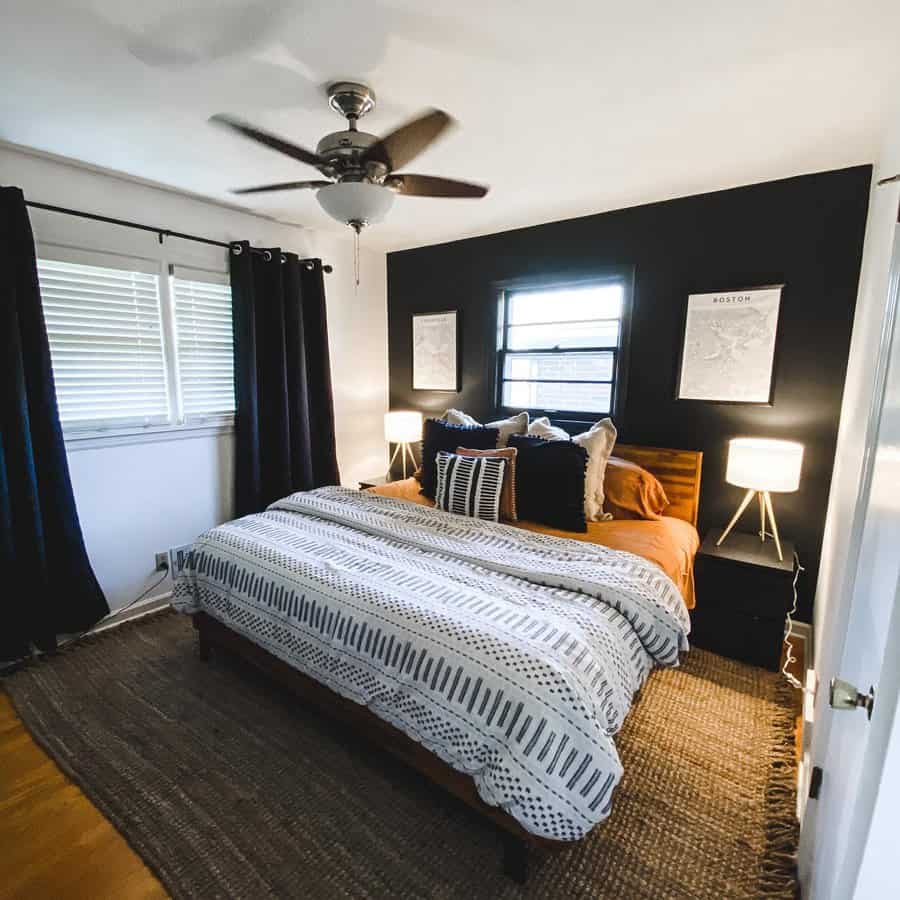 Modern bedroom with a black accent wall, striped bedding, two bedside tables with lamps, and a ceiling fan bathed in natural light from windows