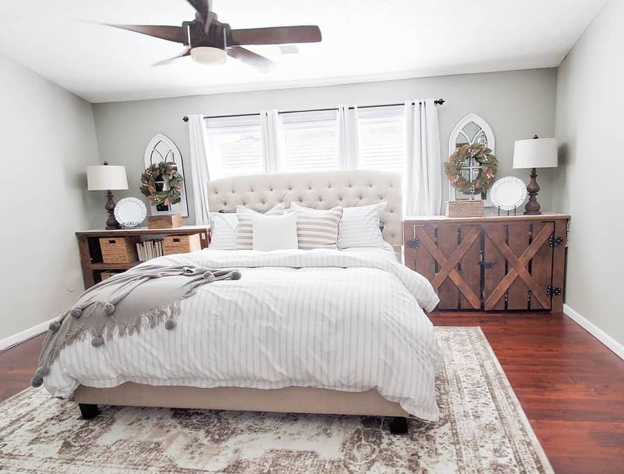 Bedroom with neutral tones featuring a tufted bed, white and gray bedding, wooden side tables, a ceiling fan, and natural light from windows