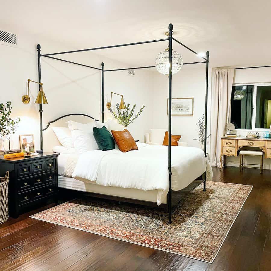 Master bedroom with a canopy bed, white bedding, decorative pillows, bedside tables, plants, a rug, and a vintage-style dressing table