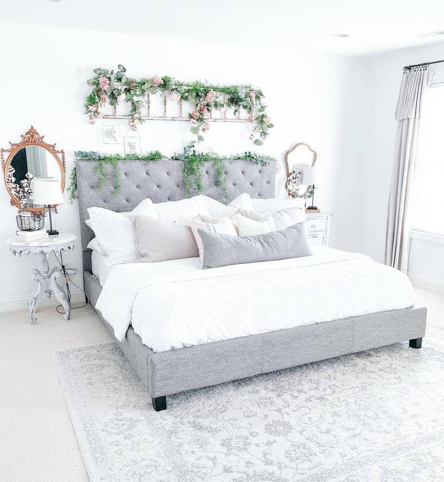 Bright bedroom with a gray tufted bed, white bedding, floral decor above the headboard, and vintage mirrors on side tables in a light-filled space