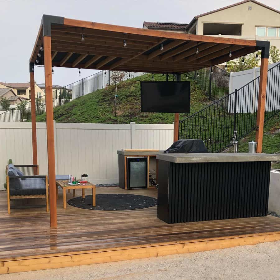 Outdoor patio with a wooden pergola, TV, sofa, table, mini fridge, and grill on a wooden deck; surrounded by a fence and houses