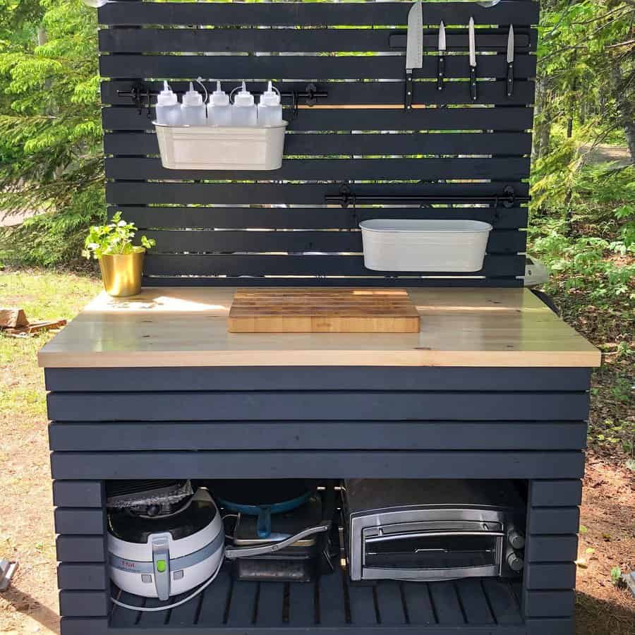 Outdoor kitchen setup with a wooden counter, hanging kitchen tools, spice bottles, and storage below, surrounded by greenery