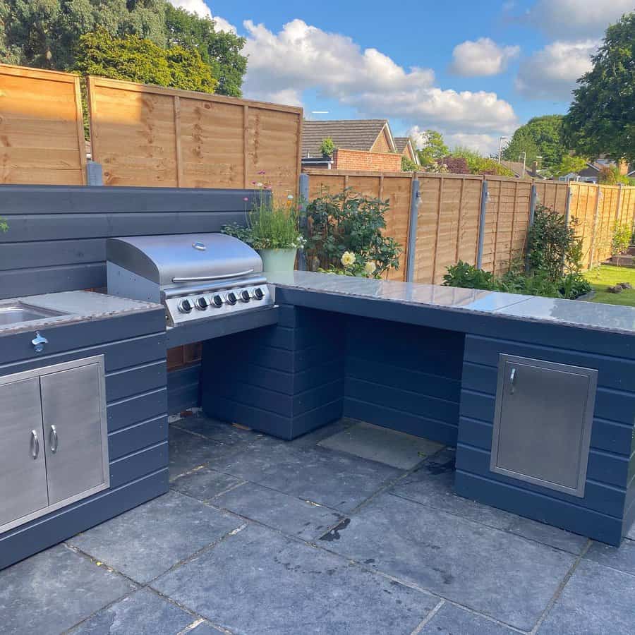 Outdoor kitchen with a stainless steel grill, blue cabinets, and a tiled countertop under a partly cloudy sky; wooden fence in the background