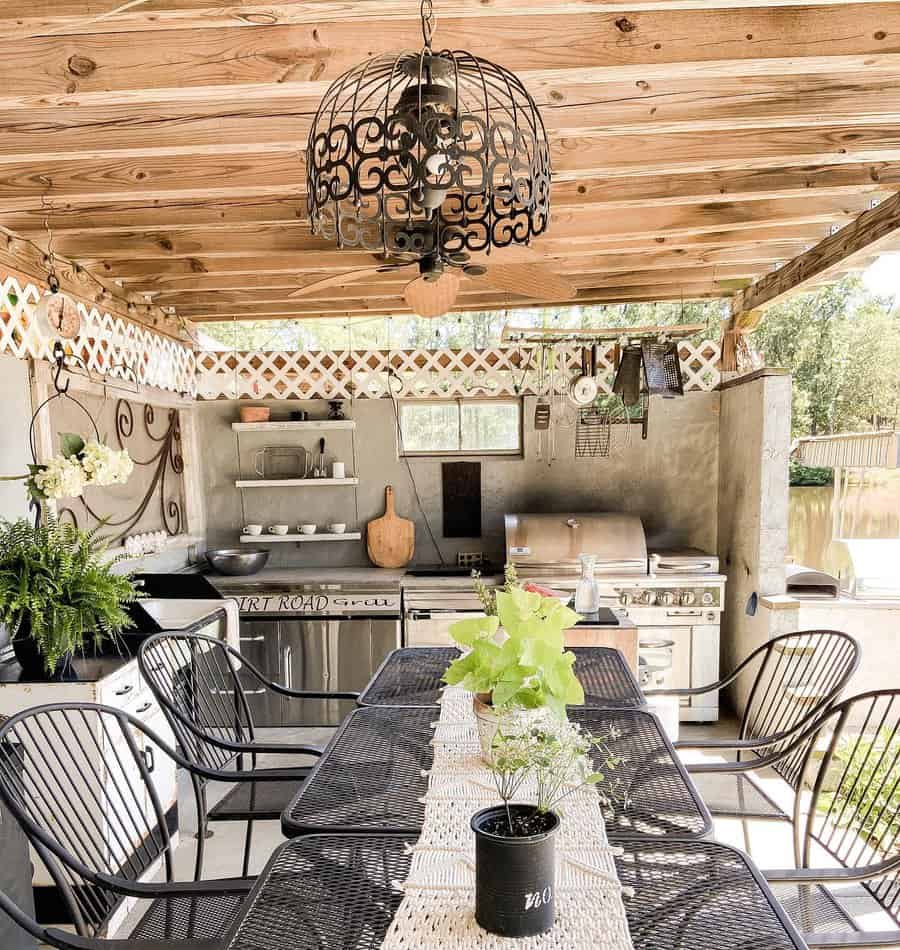 An outdoor kitchen with a wooden pergola, grill, oven, and black chairs around a table decorated with plants