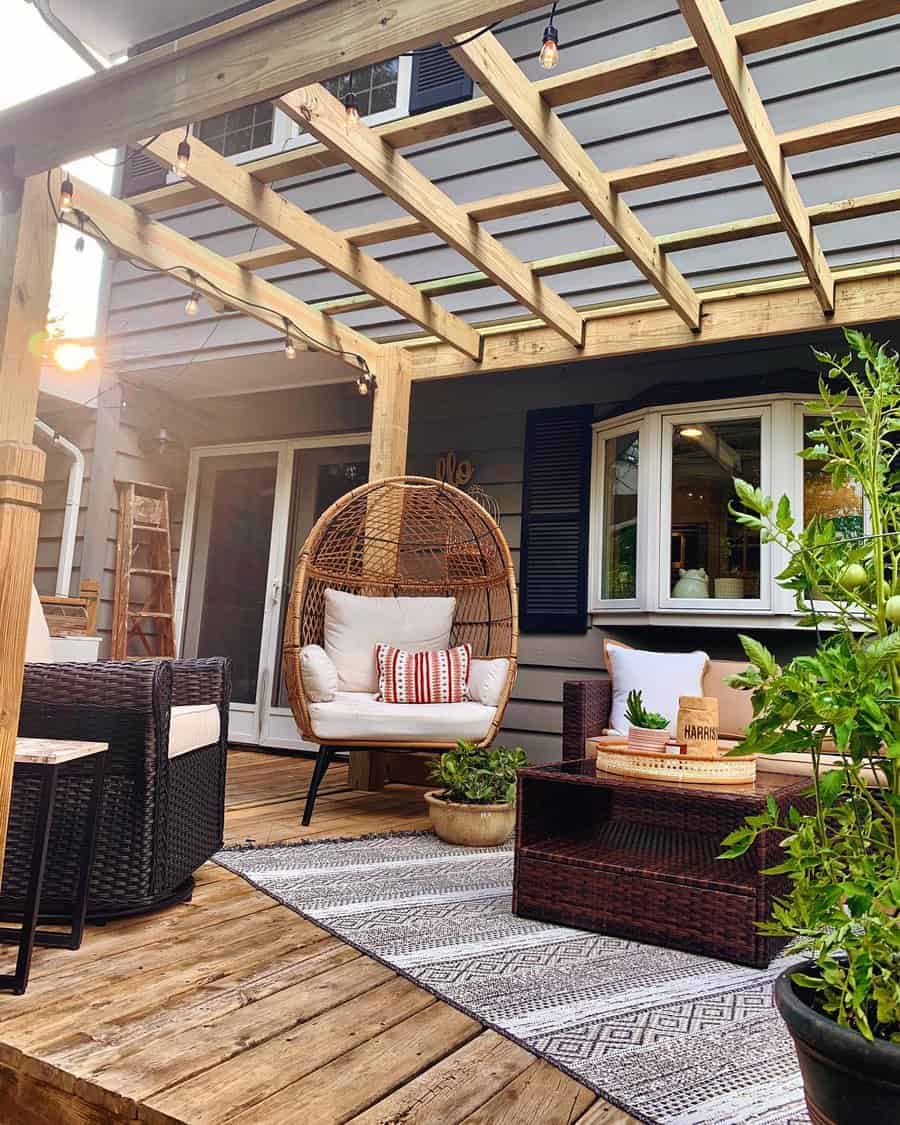 A cozy patio with a wooden pergola, hanging chair, cushioned seating, a patterned rug, and potted plants on wooden decking