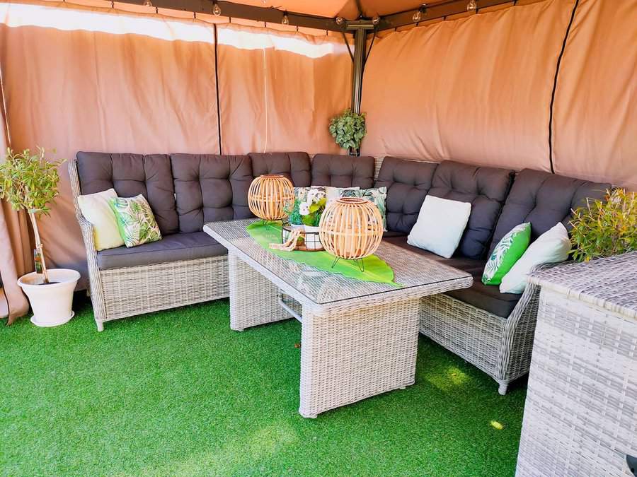 Outdoor corner sofa with gray cushions, wicker table, decorative lanterns, green throw pillows, and potted plants on artificial grass