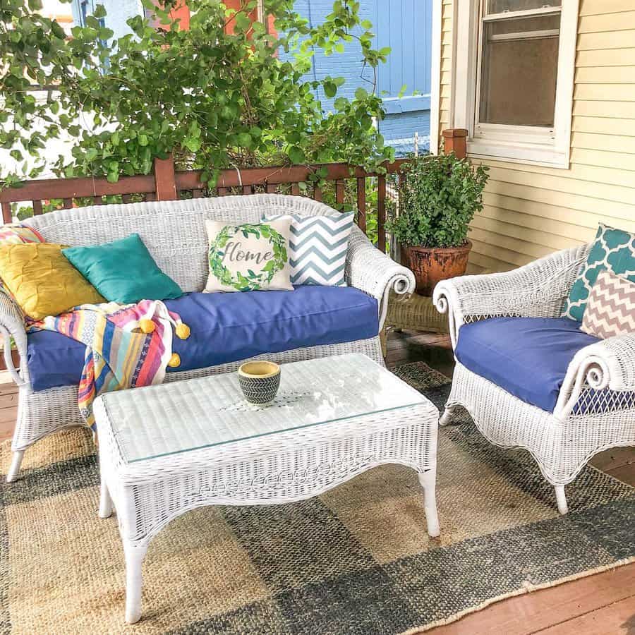 Cozy porch with white wicker furniture, blue cushions, colorful pillows, and a coffee cup on a glass-top table; lush greenery in background