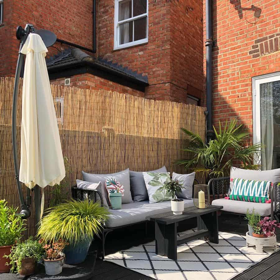 Cozy patio with plants, a white umbrella, and a brick wall backdrop. Outdoor seating with cushions around a black coffee table on a rug.