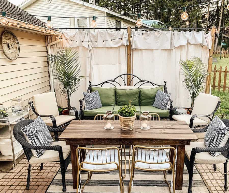 Cozy outdoor patio with a wooden table, chairs, cushioned bench, string lights, and potted plants; cream curtains create a cozy atmosphere