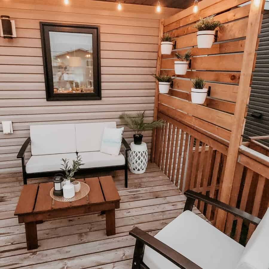 Cozy deck with white cushioned seating, wooden table, string lights, potted plants on walls, and framed picture