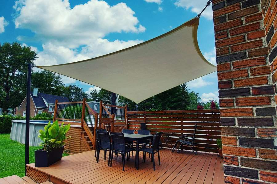 Patio with a triangular sunshade, a table with chairs, and a wooden deck, set in a green backyard under a blue sky with clouds